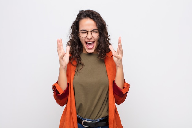 Jonge mooie vrouw die woedend schreeuwt, zich gestrest en geïrriteerd voelt met handen omhoog in de lucht en zegt waarom ik tegen de witte muur