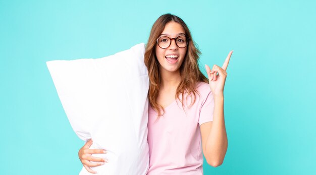 Jonge mooie vrouw die zich een gelukkig en opgewonden genie voelt na het realiseren van een idee dat ze een pyjama draagt en een kussen vasthoudt