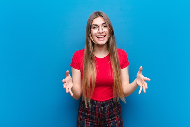 Jonge mooie vrouw die zich gelukkig, verbaasd, gelukkig en verrast voelt, zoals omg serieus zegt? Ongelofelijk tegen blauwe muur