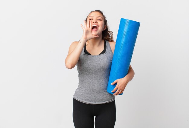 Jonge mooie vrouw die zich gelukkig voelt, een grote schreeuw geeft met de handen naast de mond en een yogamat vasthoudt