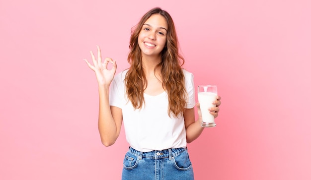 Jonge mooie vrouw die zich gelukkig voelt, goedkeuring toont met een goed gebaar en een glas melk vasthoudt