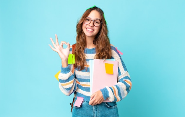 Jonge mooie vrouw die zich gelukkig voelt, goedkeuring toont met een goed gebaar met een tas en boeken vasthoudt