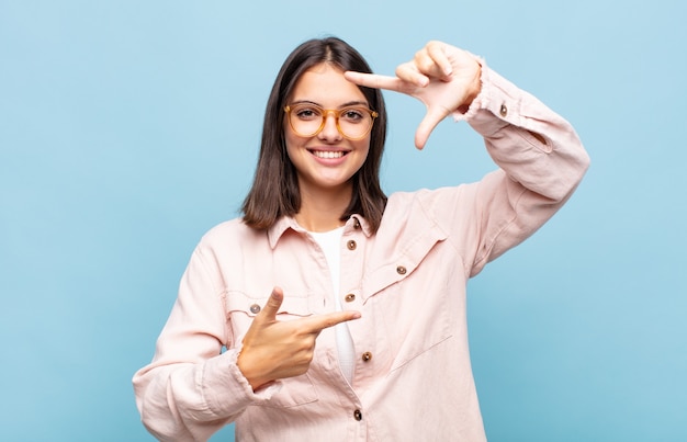 Jonge mooie vrouw die zich gelukkig, vriendelijk en positief voelt, lacht en een portret of fotolijst met handen maakt