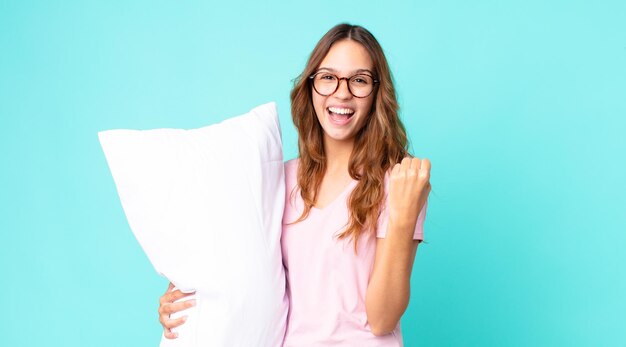 Jonge mooie vrouw die zich geschokt voelt, lacht en succes viert met een pyjama en een kussen vast
