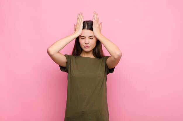 Jonge mooie vrouw die zich gestrest en angstig, depressief en gefrustreerd voelt met hoofdpijn, beide handen opheffend om over de roze muur te gaan