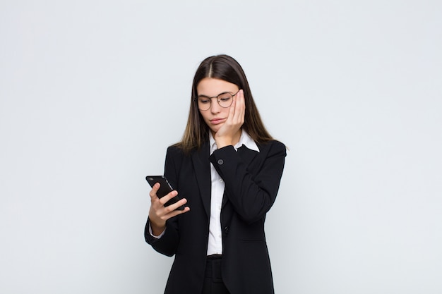 Jonge mooie vrouw die zich verveeld, gefrustreerd en slaperig voelt na een vermoeiende, saaie en vervelende taak, die haar gezicht met de hand met een mobiele telefoon vasthoudt