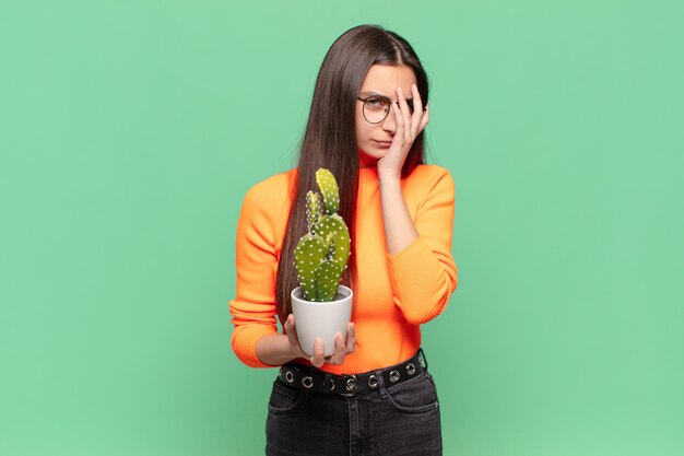 Jonge mooie vrouw die zich verveeld, gefrustreerd en slaperig voelt na een vermoeiende, saaie en vervelende taak, gezicht met de hand vasthoudend. cactusconcept