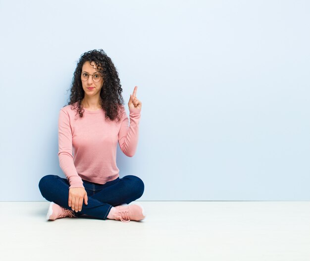 Jonge mooie vrouw die zich voelt als een genie die vinger trots in de lucht houdt na het realiseren van een geweldig idee, zeggende eureka zittend op de vloer