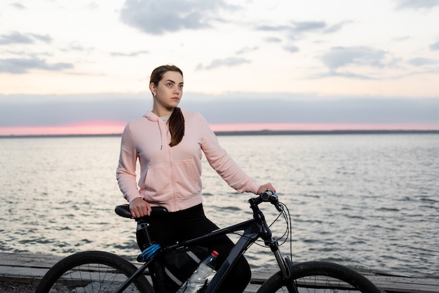 Jonge mooie vrouw, een atleet met een fiets, kijkt vanaf de vroege ochtend naar de dageraad over de zee en rust uit van een fietstocht.