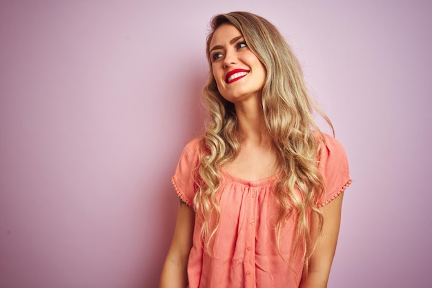 Jonge, mooie vrouw, gekleed in een t-shirt die over een roze geïsoleerde achtergrond staat, wegkijkend naar de andere kant met een glimlach op het gezicht, natuurlijke uitdrukking Lachend zelfverzekerd