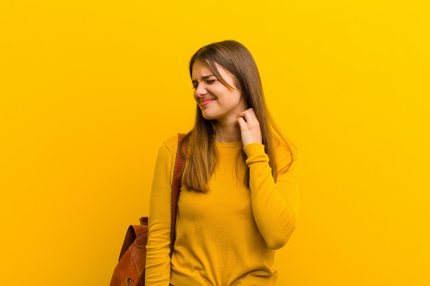 Jonge mooie vrouw gevoel gestrest, gefrustreerd en moe, wrijven pijnlijke nek, met een bezorgd, onrustige blik tegen oranje achtergrond