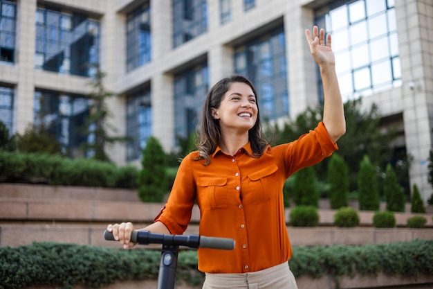 Jonge mooie vrouw glimlacht en rijdt op een elektrische scooter om langs kantoorgebouwen te werken Ecologisch transportconcept