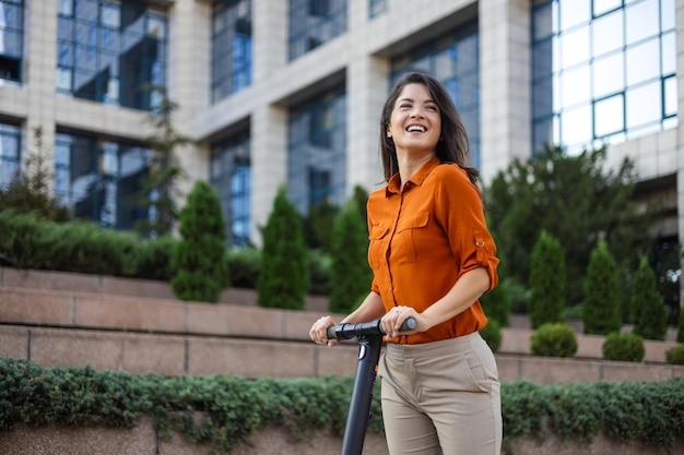 Jonge mooie vrouw glimlacht en rijdt op een elektrische scooter om langs kantoorgebouwen te werken Ecologisch transportconcept