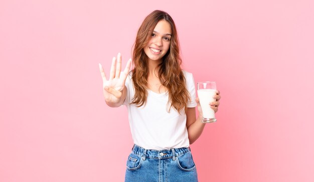 jonge mooie vrouw glimlacht en ziet er vriendelijk uit, toont nummer vier en houdt een glas melk vast