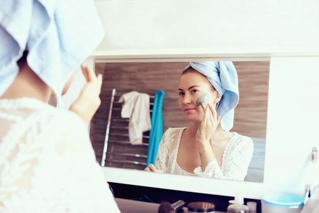 Foto jonge mooie vrouw in badjas en met handdoek op hoofd die gezichtsmasker voor spiegel in badkamers verwijderen. huidverzorging en schoonheid concept