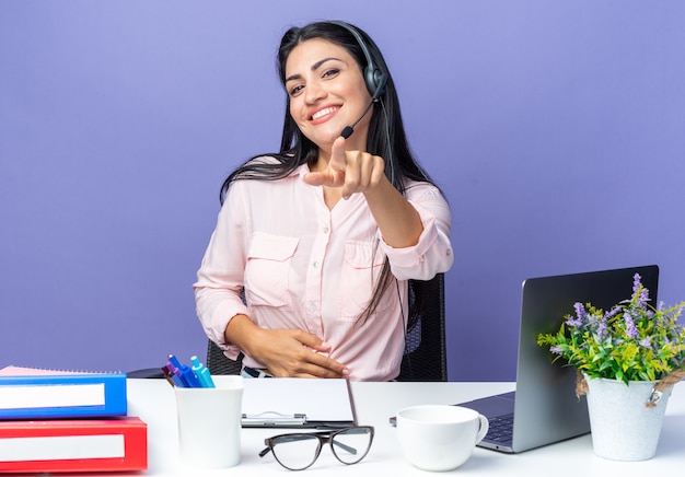 Jonge mooie vrouw in casual kleding met koptelefoon en microfoon wijzend met wijsvinger ay camera glimlachend zelfverzekerd zittend aan de tafel met laptop over blauwe muur werken op kantoor