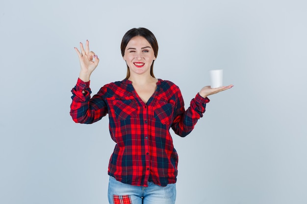 Jonge mooie vrouw in casual shirt, jeans met plastic glas, ok teken tonen en gelukzalig kijken, vooraanzicht.