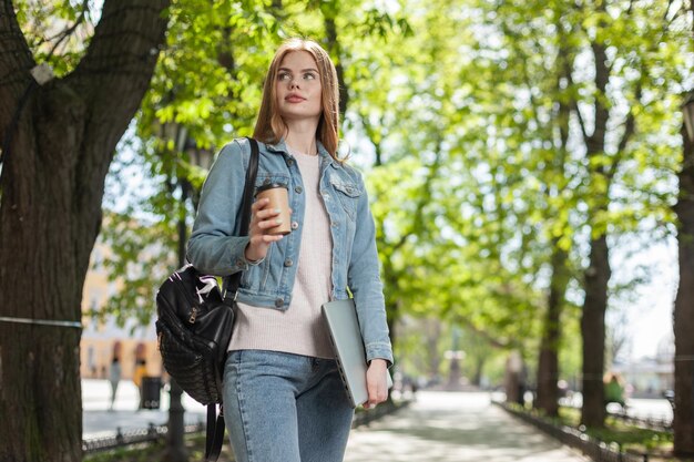 Jonge mooie vrouw in een spijkerjasje houdt een laptop en een kopje koffie in de stad