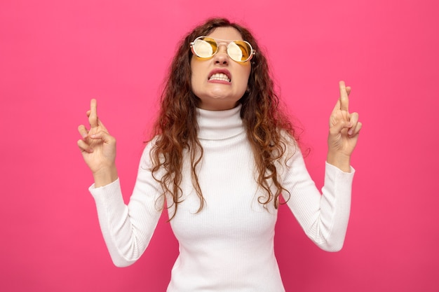 Jonge, mooie vrouw in een witte coltrui met een gele bril die er verward uitziet met een geïrriteerde uitdrukking die de vingers kruist en een wens doet die op roze staat