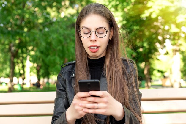 Jonge mooie vrouw in een zwart leren jas schrijft een bericht of speelt games of bekijkt video's of social media nieuws aan de telefoon zittend op een bankje in een prachtig groen zomer- of lentepark