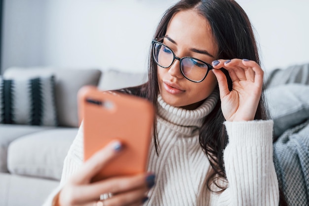 Jonge mooie vrouw in glazen om alleen thuis te zitten met telefoon in handen.