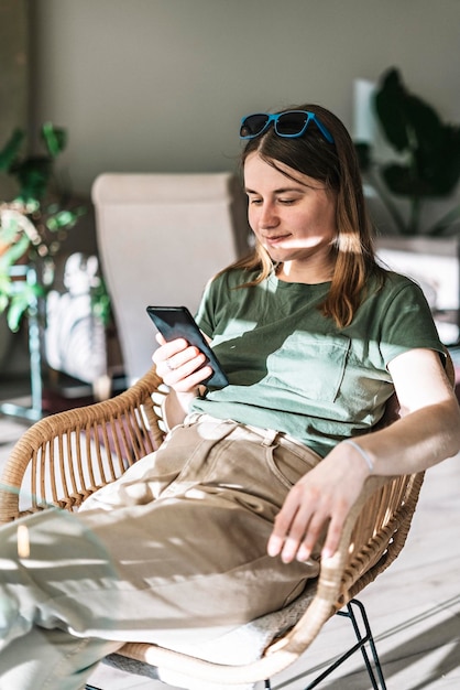 Jonge mooie vrouw in groene beige kleren zit in een rieten stoel in het zonlicht met behulp van een smartphone