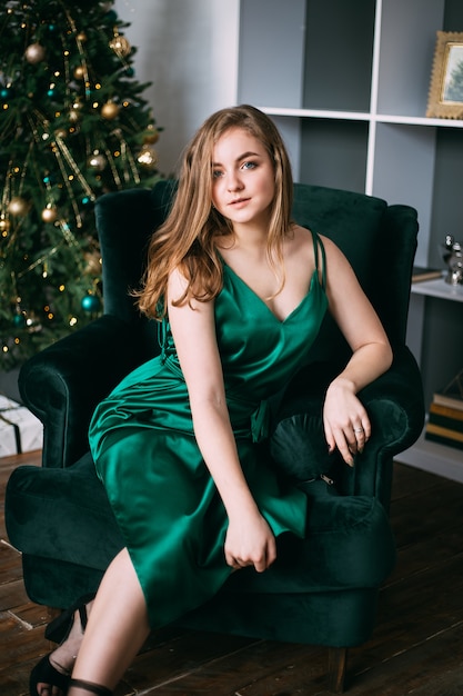 Jonge mooie vrouw in groene elegante avondjurkzitting op leunstoel dichtbij kerstmisboom, nieuw jaar