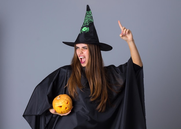 Jonge mooie vrouw in kostuum van heks met zwarte hoed die een pompoen op blauwe achtergrond in studio houdt