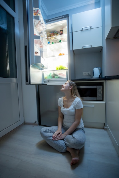 Jonge mooie vrouw in pyjama zittend op de keukenvloer en kijken naar open koelkast