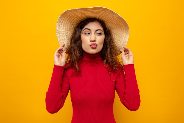 Jonge, mooie vrouw in rode coltrui in zomerhoed die opzij kijkt en haar lippen houdt alsof ze over een oranje muur gaat kussen