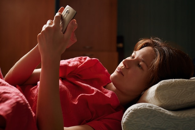 Foto jonge mooie vrouw in rode kleding die op het bed liggen en smartphone gebruiken bij avond