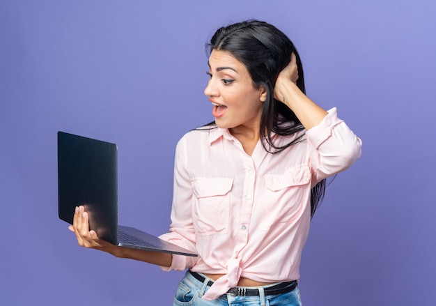 Jonge, mooie vrouw in vrijetijdskleding die een laptop vasthoudt en naar het scherm kijkt verbaasd en verrast over een blauwe muur