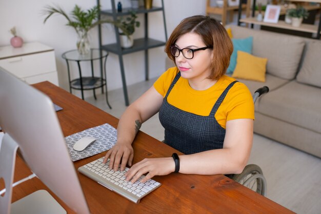 Jonge mooie vrouw in vrijetijdskleding zittend in een rolstoel door bureau en kijken naar computerscherm tijdens het leren op afstand van huis