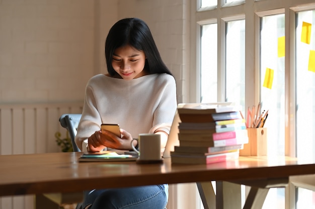 Jonge mooie vrouw in wit katoenen overhemd die / smartphone in haar handen houden gebruiken terwijl samen het zitten met laptop, koffiekop, potloodhouder en stapel boeken die op houten lijst zetten.