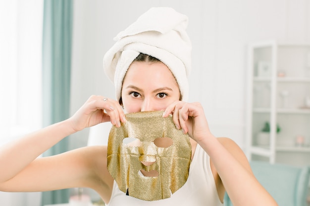 Jonge mooie vrouw in witte handdoek na bad met papier gouden gezichtsmasker. Vrouw die gouden gezichtsmasker krijgt.