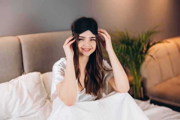 Jonge mooie vrouw in witte zijden pyjama met een zwart slaapmasker op haar hoofd. een brunette met een slaapmasker op haar hoofd zit op een bed en lacht. ochtend of avond in het bed in de slaapkamer.