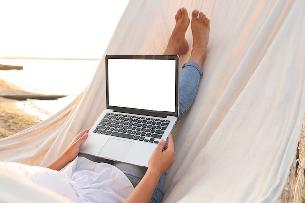 Jonge mooie vrouw liggend in een hangmat met een laptop in het resort. Bespotten, leeg wit scherm.