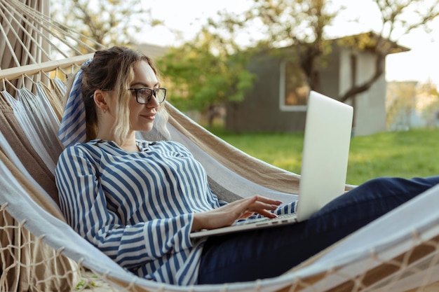 Jonge mooie vrouw ligt in een hangmat met een laptop in een bril buiten thuis