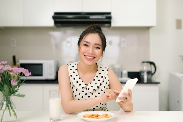 Jonge mooie vrouw met behulp van slimme telefoon zitten aan de tafel in de keuken