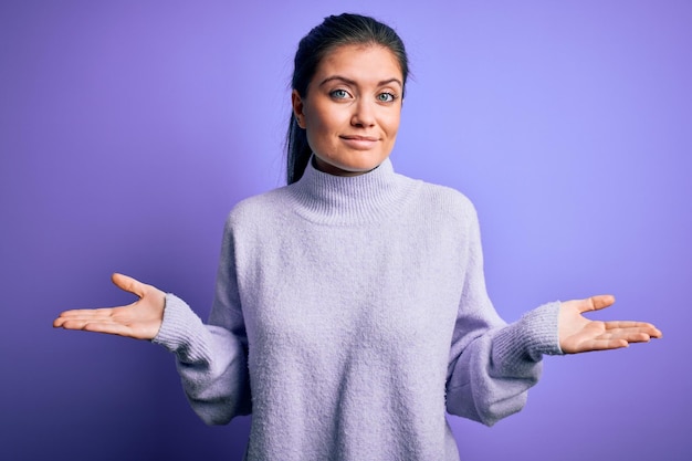 Jonge mooie vrouw met blauwe ogen dragen casual coltrui over roze achtergrond clueless en verwarde uitdrukking met opgeheven armen en handen twijfel concept