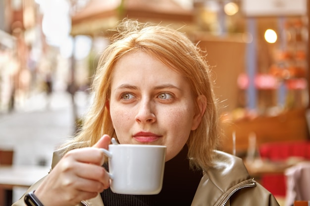 Jonge mooie vrouw met blond haar zit buiten café met een klein kopje koffie in haar handen