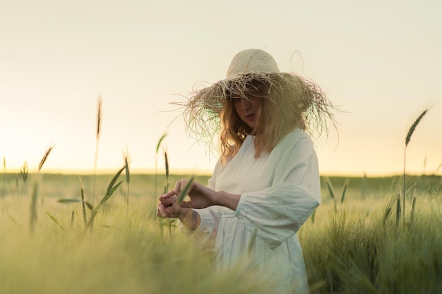jonge mooie vrouw met blond lang haar in een witte jurk in een strohoed verzamelt bloemen op een tarweveld. Vliegende haren in de zon, zomer. Tijd voor dromers, gouden zonsondergang.