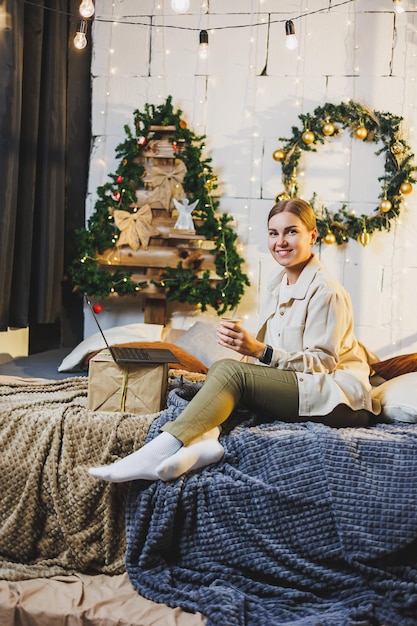 Jonge mooie vrouw met blond lang haar in knusse kleding met koffie in handen met behulp van laptop film kijken en glimlachend op bed in kamer met kerstboom thuis nieuwjaarsvakantie