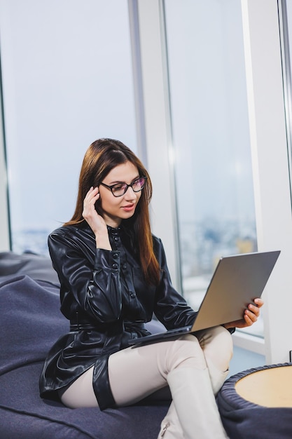Jonge mooie vrouw met een bril werkt op een laptop terwijl ze in een moderne werkruimte zit Freelance werken op afstand