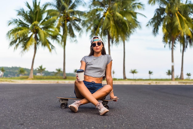 Jonge mooie vrouw met een pet die op een skateboard zit en koffie drinkt