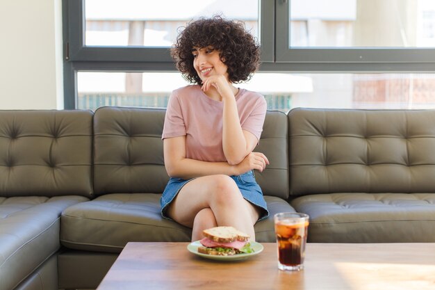 Jonge mooie vrouw met een sandwich zittend op een bank thuis