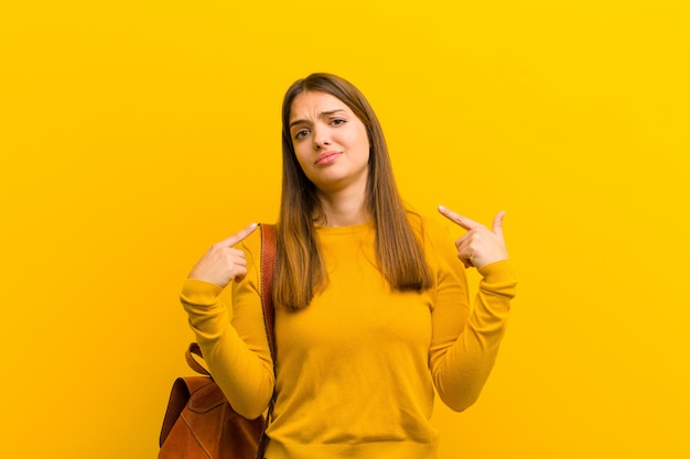 Jonge mooie vrouw met een slechte houding die trots en agressief kijkt, naar omhoog richt of pretteken met handen tegen oranje achtergrond maakt