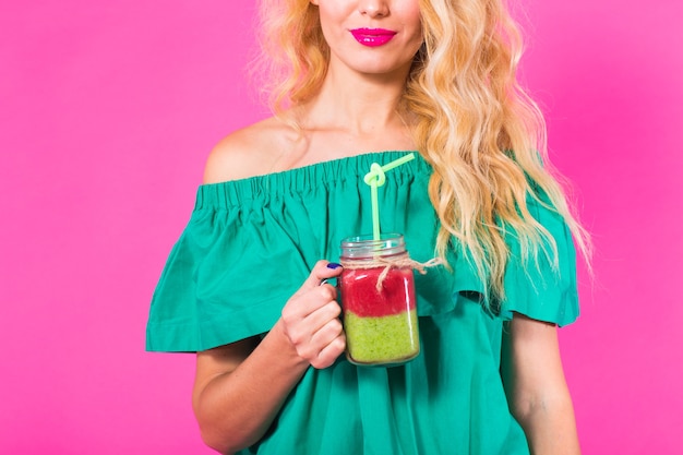 Jonge mooie vrouw met groene smoothie op roze achtergrond.