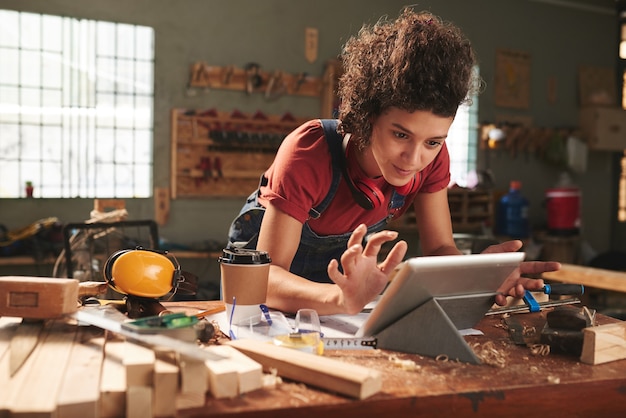 Jonge mooie vrouw met krullend haar kijken naar houtbewerking tutorial op digitale tablet terwijl leunend op rommelige tafel bedekt met zaagsel