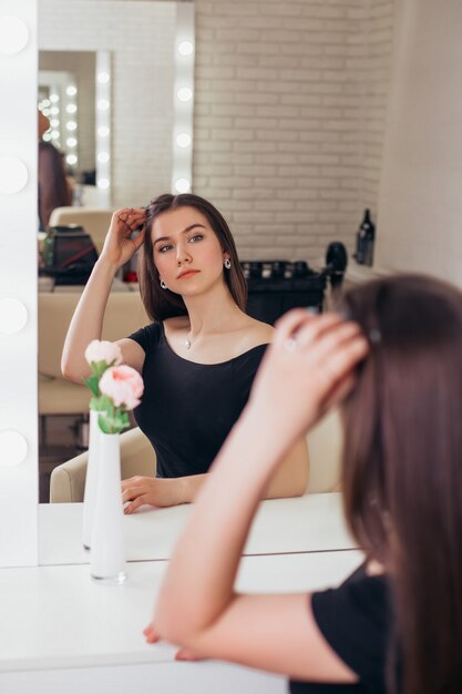 Jonge mooie vrouw met lang gezond donkerbruin haar die de spiegel in kapsalon bekijken